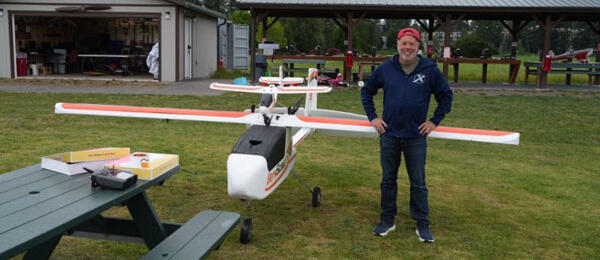Man standing next to a large model airplane on grass, with hangar in the background.