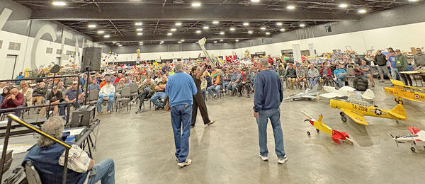 Crowded hall with people sitting and standing, facing a stage with small aircraft models.
