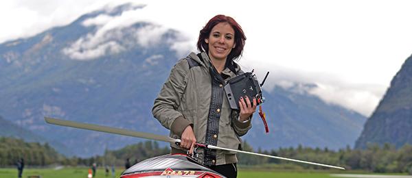Person holding a remote control beside a red model helicopter in a mountainous landscape.
