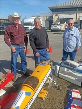 Three men stand behind model airplanes outdoors, one in a cowboy hat.