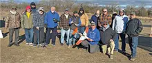 Group of 14 men outdoors, holding a model airplane. Cloudy sky and trees in the background.