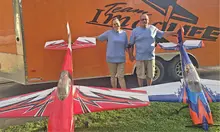 Two people in blue shirts beside large model planes and an orange trailer.