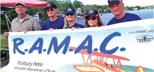 Five people holding a banner for R.A.M.A.C. by a lakeside.