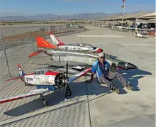 Man seated next to large model planes on a sunny airfield.