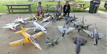 Three people seated behind an array of model jets on concrete.