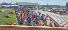 Crowd gathered outdoors near a fence, with tents and small airplanes nearby.