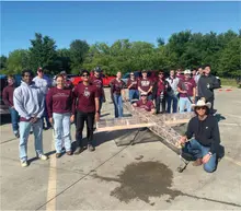 Group of people standing around a large model airplane outdoors, wearing maroon shirts.
