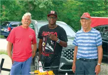 Three men standing in front of parked cars, one wearing a NASA T-shirt and cap.