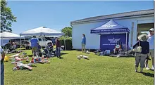 People gather near model planes under tents on a sunny lawn.