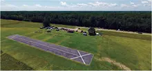 Aerial view of a small airstrip with parked cars and tents nearby, surrounded by lush greenery.