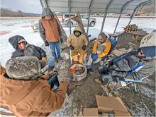 Six people in winter clothes sit around a small fire under a shelter, with snow in the background.