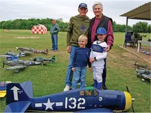 Four people smiling by model airplanes on grass. Child in baseball uniform.