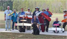 Group of nine people holding model airplanes on a grassy field; one person with a black dog.