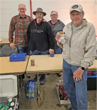 Four men pose indoors by tables, one holding a snack, with drinks and a cooler nearby.