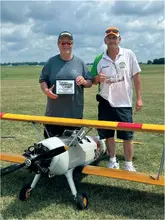 Two men standing with a model plane; one holds a "NATS 2023" sign.