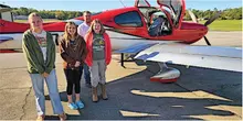 Four people stand smiling in front of a red and white small aircraft on a sunny day.