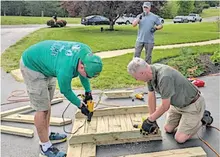 Two men assembling wooden boards with drills, a third man stands nearby on a driveway.
