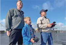 Two men and a boy control a drone outdoors, sky in the background.