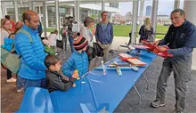 Man shows model airplanes to family at indoor event; blue table, diverse crowd.
