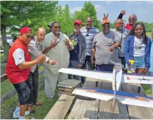 Group of people outdoors smiling beside a model airplane on a picnic table.