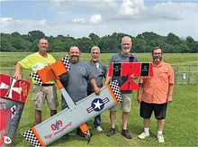 Five men holding model airplanes on a grassy field, smiling at the camera.