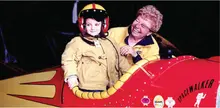 Boy in flight suit and helmet with cheerful woman beside red plane labeled "Spacewalker".