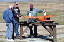 Three men examining an orange model airplane on a stand in a field.