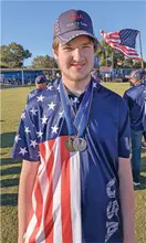 Young man in USA-themed shirt and cap with medals, standing outdoors, American flag in background.