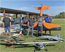 Four men stand behind model airplanes on grass under a clear sky.