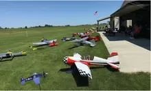 Model airplanes lined up on grass near a hangar with people seated inside.