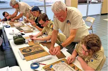 Scouts and leaders work on cardboard crafts at a table indoors.