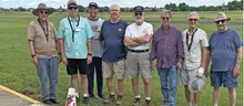 Eight men standing in a row on a grassy field wearing casual clothing and hats.