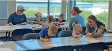 Kids and adults seated at tables coloring and writing in a bright room.
